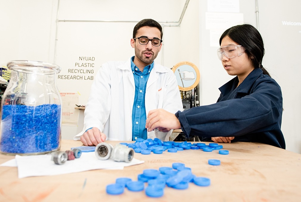 Two students working together with blue water bottle caps and other materials in the NYUAD Plastic Recycling Research Lab.