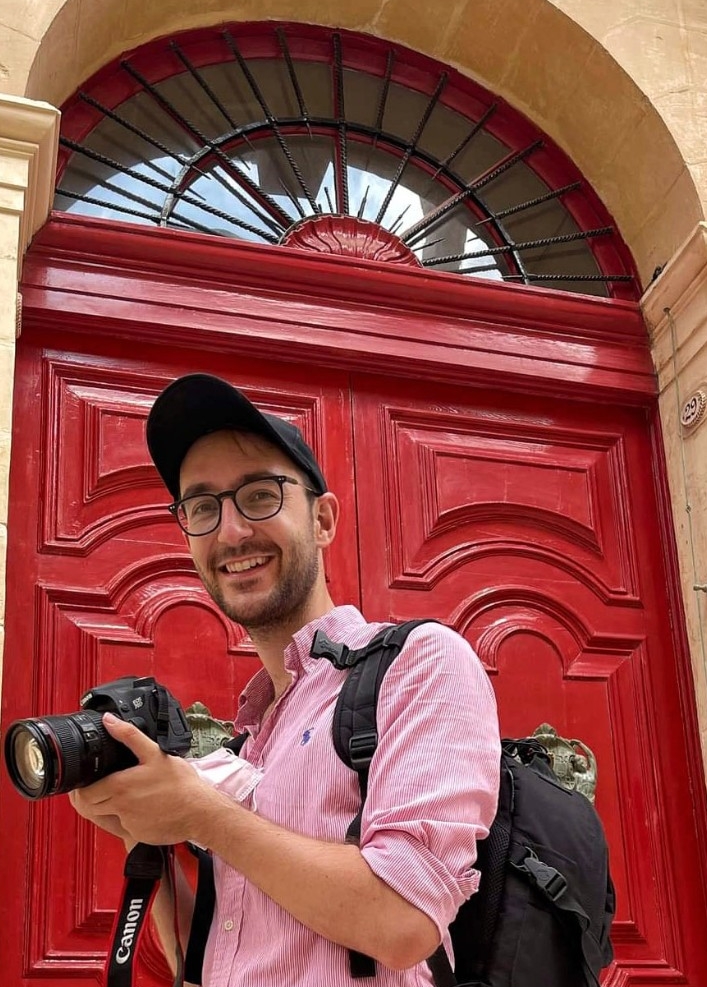 Samuel Ridgeway, NYUAD Class of 2016, hoding a camera, stands in front of a bright red door.