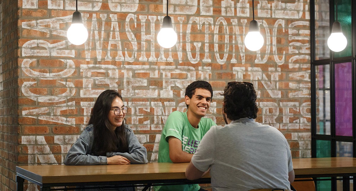 Three students are seated at a table in front of a brick wall with locations of NYU Study Away sites painted on it: Abu Dhabi, Washington DC, Accra, Berlin, etc.