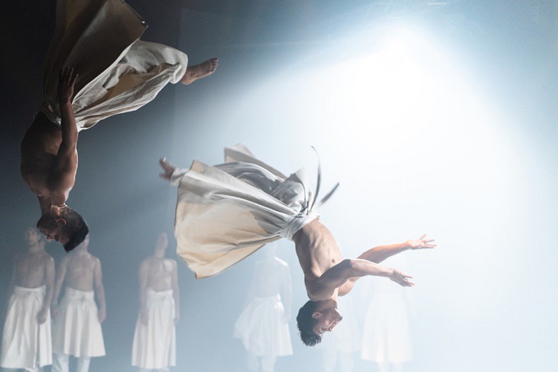 A group of dancers perform, two caught in the middle of a flip, both are in the air, upside down.