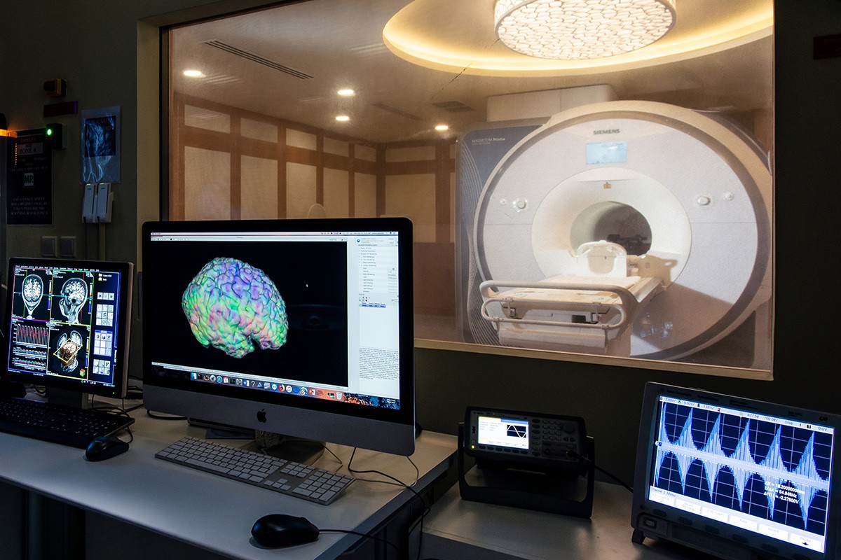 Center for Brain Health at NYU Abu Dhabi. A view of an MRI lab with brain scans on several monitors, and an MRI scanner in the background.